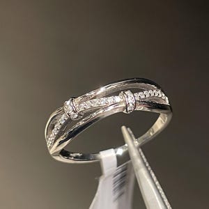 Puede incluir: Un anillo de plata con un diseño único. El anillo presenta tres bandas, con dos bandas exteriores que se curvan alrededor de una banda central adornada con pequeñas piedras transparentes. El anillo es sujetado por unas pinzas sobre un fondo neutro.