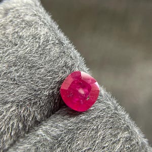 Op de afbeelding: Een gefacetteerde robijn in kussensnede. De robijn heeft een levendige, dieprode kleur met een kleine interne insluiting. De edelsteen ligt op een grijze, getextureerde achtergrond, die de glans en kleur benadrukt.