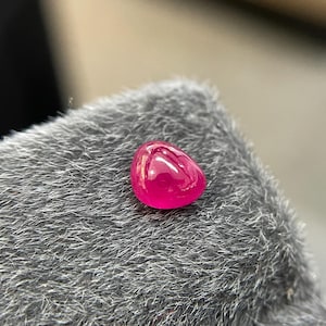 Può includere: Una gemma di rubino lucidata a forma di goccia. La gemma è di un colore rosso-rosa intenso e saturo, con una superficie liscia e riflettente. Il rubino è posto su uno sfondo grigio strutturato.