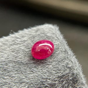 Pode incluir: Uma pedra preciosa de rubi polida, em forma oval. O rubi é de uma cor vermelha profunda e vibrante com uma superfície lisa e reflexiva. A pedra preciosa está sobre um fundo cinza texturizado.