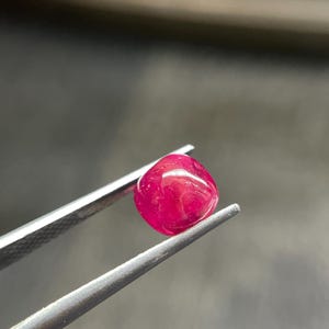 Może przedstawiać: Wypolerowany kamień szlachetny rubinu, trzymany przez srebrną pęsetę. Rubin ma głęboki, nasycony czerwony kolor, o gładkim, zaokrąglonym kształcie. Pęseta stanowi kontrastujący metaliczny element w stosunku do bogatego odcienia kamienia. Obraz jest dobrze oświetlony.