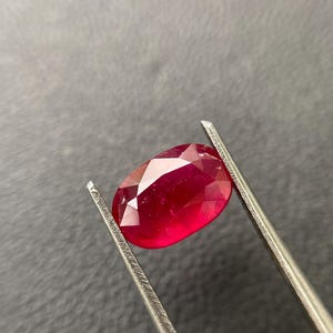 Peut inclure: Un rubis de forme ovale et facetté, maintenu par une pince argentée. La pierre précieuse est d'un rouge profond et vif, avec de multiples facettes capturant la lumière. Le fond est gris neutre.