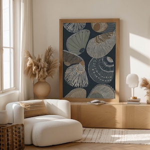 Modern coastal seashell wall art print in navy and sandy beige with hand-drawn textures, leaning on a low cabinet with sculptural vases and dried grasses.