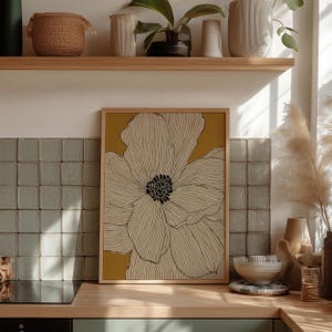 Simple botanical bloom in cream on warm yellow with black center, modern poster style, framed wall art print styled on a kitchen counter by tile.