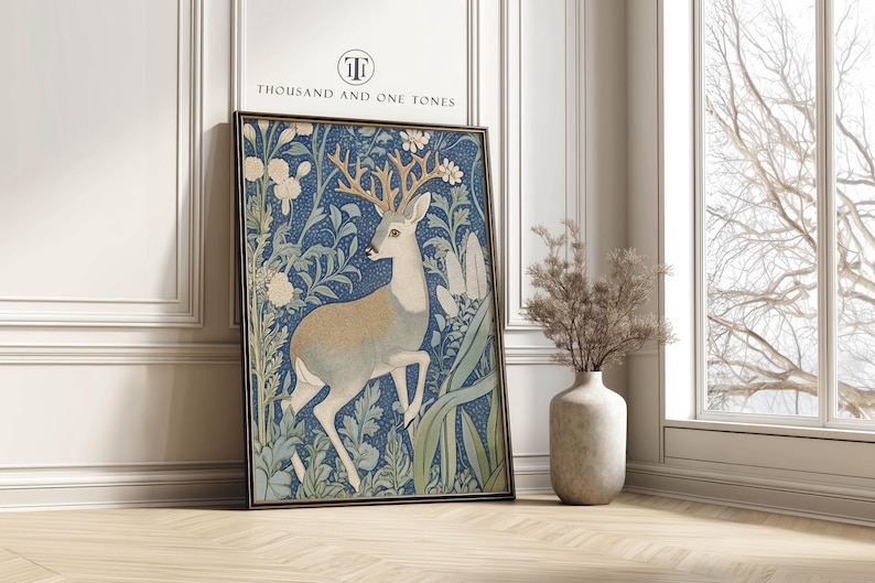 Blue and cream vintage botanical deer wall art print with intricate foliage is framed and leaning against a white paneled wall beside a tall window and ceramic vase.