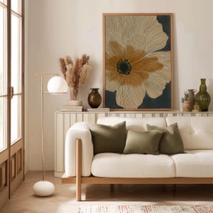 Minimalist floral poster in beige and black with delicate linework and a bold center, calm contemporary tone, framed above a console behind a sofa.