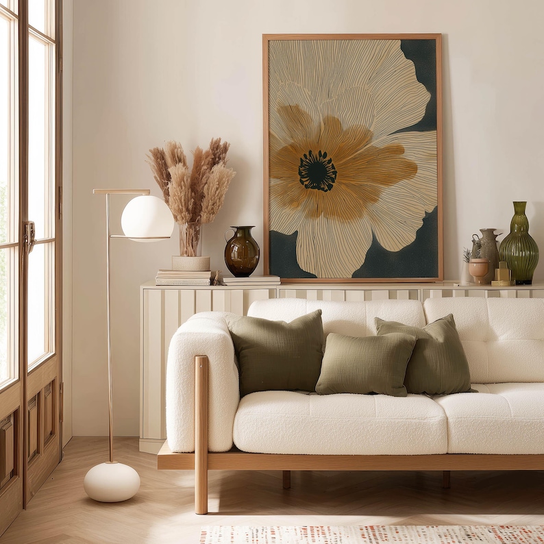 Minimalist floral poster in beige and black with delicate linework and a bold center, calm contemporary tone, framed above a console behind a sofa.