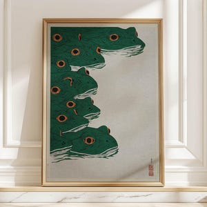 May include: A framed art print featuring a stylized illustration of several green frogs with orange eyes. The frogs are arranged in a vertical stack, with their heads and eyes visible. The artwork has a minimalist aesthetic and is set against a neutral background.