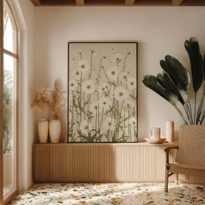 Serene botanical poster featuring white daisies and muted green foliage on beige, centered above a low cabinet with vases and greenery in a bright corner interior.