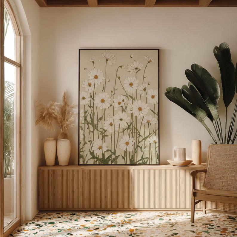 Serene botanical poster featuring white daisies and muted green foliage on beige, centered above a low cabinet with vases and greenery in a bright corner interior.