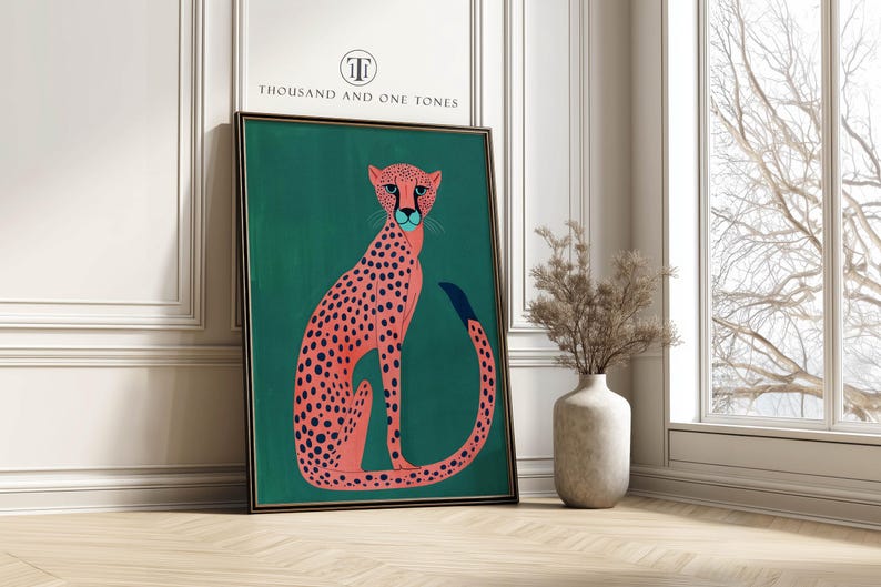 Modern art print of a pink spotted cheetah on a deep teal background with a playful modern mood, framed art print leaning against a white paneled wall by a tall window and vase of dried branches.