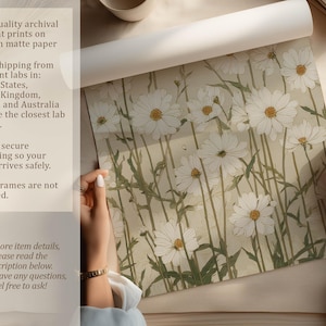 Hands unrolling a physical art print of white daisies on beige with sage green stems, photographed on a neutral tabletop beside a cup and paper-quality overlay text.
