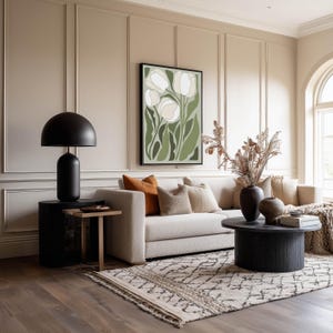 Warm floral wall art print in green and cream with an earthy, inviting mood, hanging above a beige sofa in a living room with paneled walls, a patterned rug, and round black coffee table.