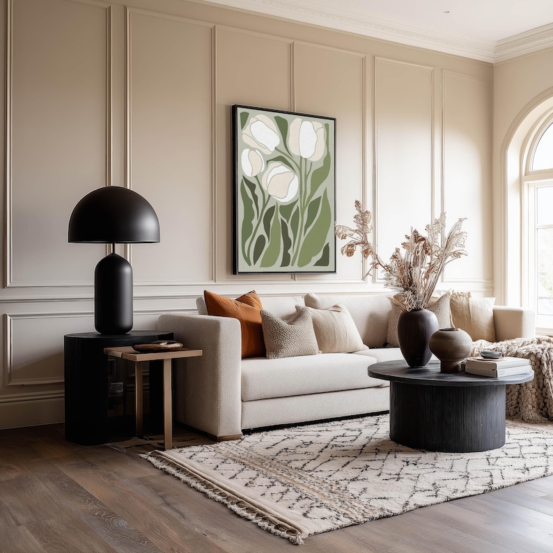 Warm floral wall art print in green and cream with an earthy, inviting mood, hanging above a beige sofa in a living room with paneled walls, a patterned rug, and round black coffee table.