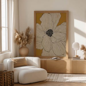 Graphic botanical flower with cream petals and a black center on mustard, modern mood, framed wall art print leaning on a low cabinet in a bright lounge.