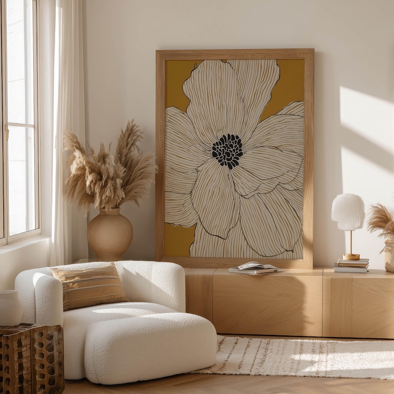 Graphic botanical flower with cream petals and a black center on mustard, modern mood, framed wall art print leaning on a low cabinet in a bright lounge.