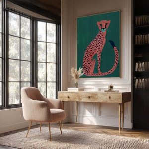 Vibrant art print of a pink cheetah with navy spots on teal, large framed above a rustic wooden desk in a bright home office with built-in bookshelves and a blush chair.