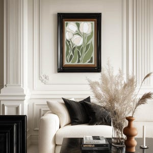 Classic botanical art print in muted greens and cream with a refined mood, framed in black and centered on an ornate paneled wall above a cream sofa styled with pampas grass.