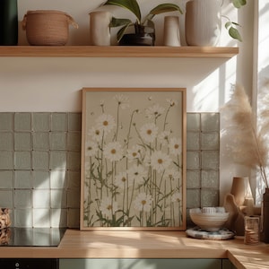 Rustic floral wall art print of white daisies with green stems on beige, framed on a kitchen counter against a tiled backsplash with bowls, candles, and a shelf.