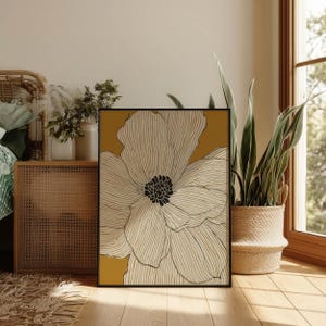 Botanical outline bloom in white and ochre with a dark center, calm modern style, framed wall art print resting on the floor in a cozy bedroom corner.