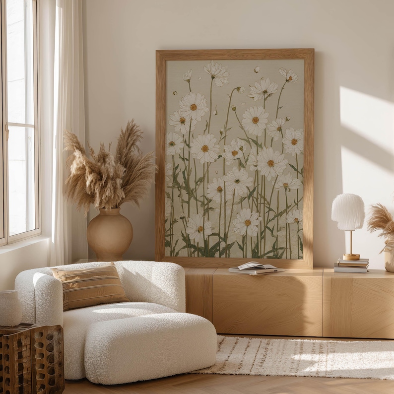 Neutral floral wall art print showing a field of white daisies with muted green stems on beige, leaning on a low cabinet with sculptural vases and dried grasses.
