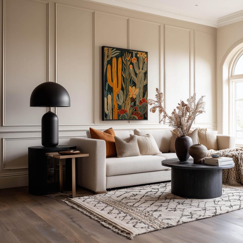 Framed desert cactus art print in black, yellow and rust tones, warm earthy mood; framed art print hanging above a beige sofa in a warm living room with paneled walls and a patterned rug.