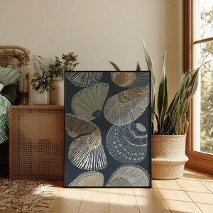 Layered blue-gray and sand seashell art print in a minimalist coastal mood, styled in a cozy bedroom corner beside a woven cabinet, plants, and a warm window.