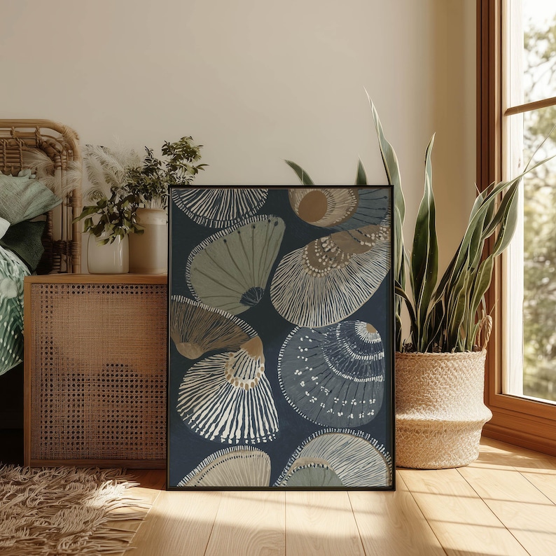Layered blue-gray and sand seashell art print in a minimalist coastal mood, styled in a cozy bedroom corner beside a woven cabinet, plants, and a warm window.