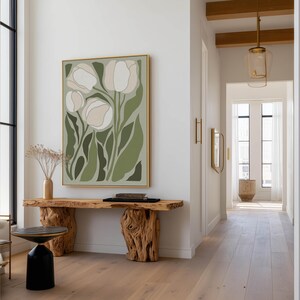 Modern floral wall art print in muted green and cream tones with a minimalist feel, hanging above a sculptural live-edge wooden console with a black accent stool and glass pendant lights.