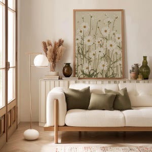 Minimal botanical poster of white daisy blossoms and thin green stems on a beige ground, displayed above a slim console with a lamp, ceramics, and green cushions.