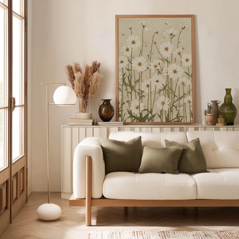Minimal botanical poster of white daisy blossoms and thin green stems on a beige ground, displayed above a slim console with a lamp, ceramics, and green cushions.