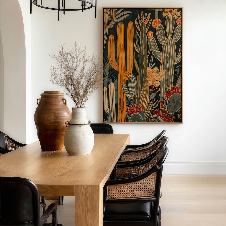 Bold desert botanical poster in black and mustard with muted teal details, contemporary mood; large framed art print hanging above a light wood dining table with cane-back chairs and ceramic vases.