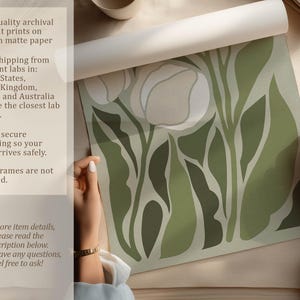 Hand-rolled tulip art print in soft sage and cream shown on a neutral tabletop being unrolled by two hands, with overlay text about archival pigment printing and matte paper.