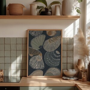 Coastal shell wall art print in deep blue with beige and gray shells, framed on a kitchen counter against tile with bowls, candles, and ceramic shelf decor.