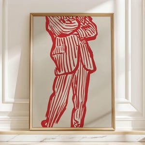 May include: A framed art print depicting a red and white striped figure. The artwork is a minimalist illustration of a person in a suit, with bold lines and a neutral background. The frame is gold.