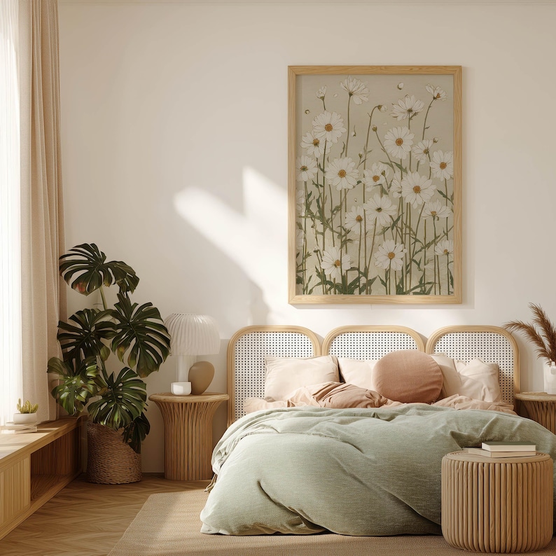 Vintage-style botanical art print of tall white daisies with green leaves over a beige background, hanging above a cane headboard with green bedding in soft daylight.