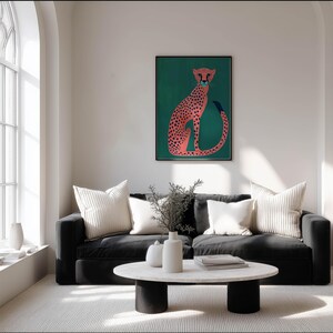 Whimsical poster of a pink cheetah on deep teal with bold navy spots, displayed on a soft white wall in a bright living room with a black sofa and arched window.