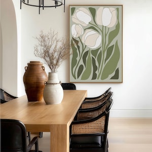 Elegant botanical poster in muted green and cream with a serene mood, large framed poster hanging above a light wood dining table with cane-back chairs and ceramic vases.