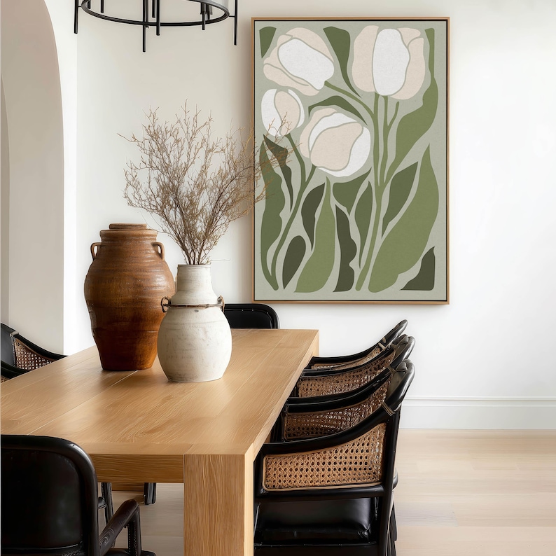 Elegant botanical poster in muted green and cream with a serene mood, large framed poster hanging above a light wood dining table with cane-back chairs and ceramic vases.
