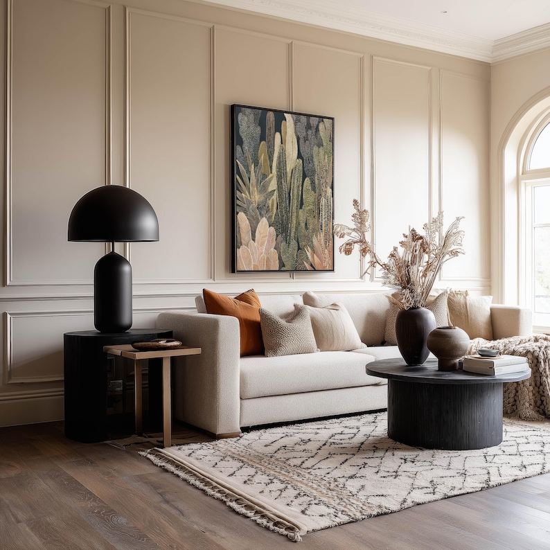 Botanical cactus scene in warm neutrals and dark background, framed art print hanging above a beige sofa with paneled walls and an earthy patterned rug.
