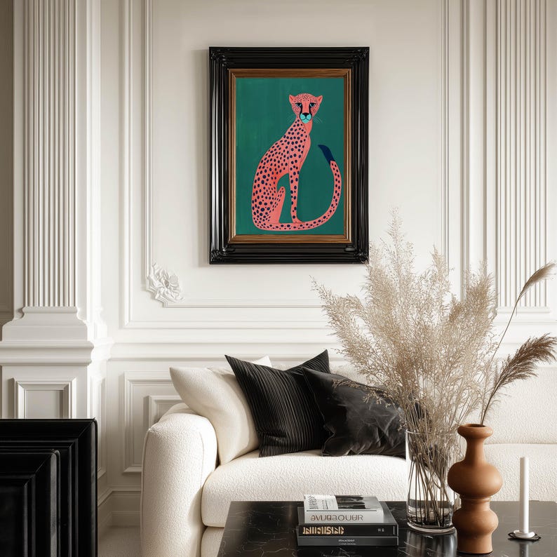 Contemporary art print of a pink spotted cheetah on deep green, centered in a classic black frame above a cream sofa styled with pampas grass and warm ceramic vases.