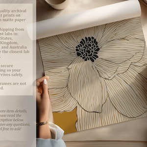 Unrolled art print showing a white linework flower on mustard yellow, crisp modern style, photographed on a tabletop with hands and paper details visible.