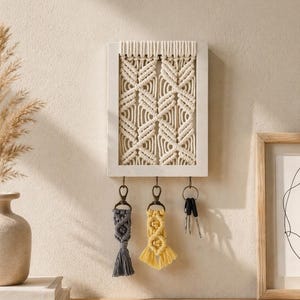 Puede incluir: Un llavero de macramé blanco con un diseño geométrico, enmarcado en un rectángulo de madera blanca. Cuelgan tres llaveros debajo: un llavero de macramé gris con borla, un llavero de macramé amarillo y un juego de llaves.