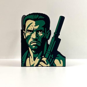 May include: A green and black pop art style image of a man holding a rifle. The man's face is shaded in green and tan, with a stern expression. The rifle is also green and black. The image is a stylized representation of a soldier.