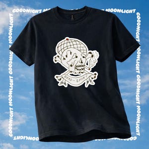 May include: A black short-sleeve t-shirt featuring a graphic of a skull wearing a checkered cap, smoking a cigarette, and positioned above crossed bones. A banner below the skull contains the text DEAD-SPORT in black capital letters.