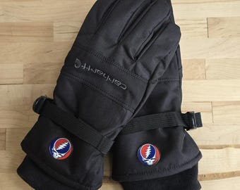 May include: Black Carhartt winter gloves with a black strap and buckle closure. The gloves have a black cuff and a colorful Steal Your Face patch. The Carhartt logo is embroidered on the top of the gloves.