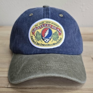 Puede incluir: Una gorra de béisbol de mezclilla azul con visera verde oliva. El frente presenta un parche circular con fondo amarillo, un logotipo de calavera con un rayo y el texto Grateful Dead, The music never stopped, Made in San Francisco since 1965.