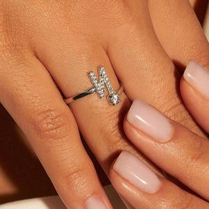 以下が含まれることがあります： 小さな輝く宝石がちりばめられた、文字「M」が特徴のシルバーリング。リングは繊細なバンドと、文字の根元に1つの大きな宝石が付いています。マニキュアされた爪の手でリングが示されています。