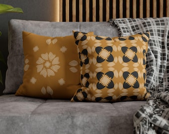 May include: Two decorative throw pillows with mustard yellow backgrounds. One pillow features a floral design with white accents, while the other has a repeating pattern of black and beige shapes. The pillows rest on a gray sofa.