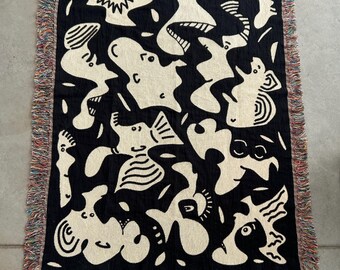 May include: Black throw blanket with a cream-colored abstract design. The blanket has a fringed edge with a mix of colors. The design features various shapes and figures.